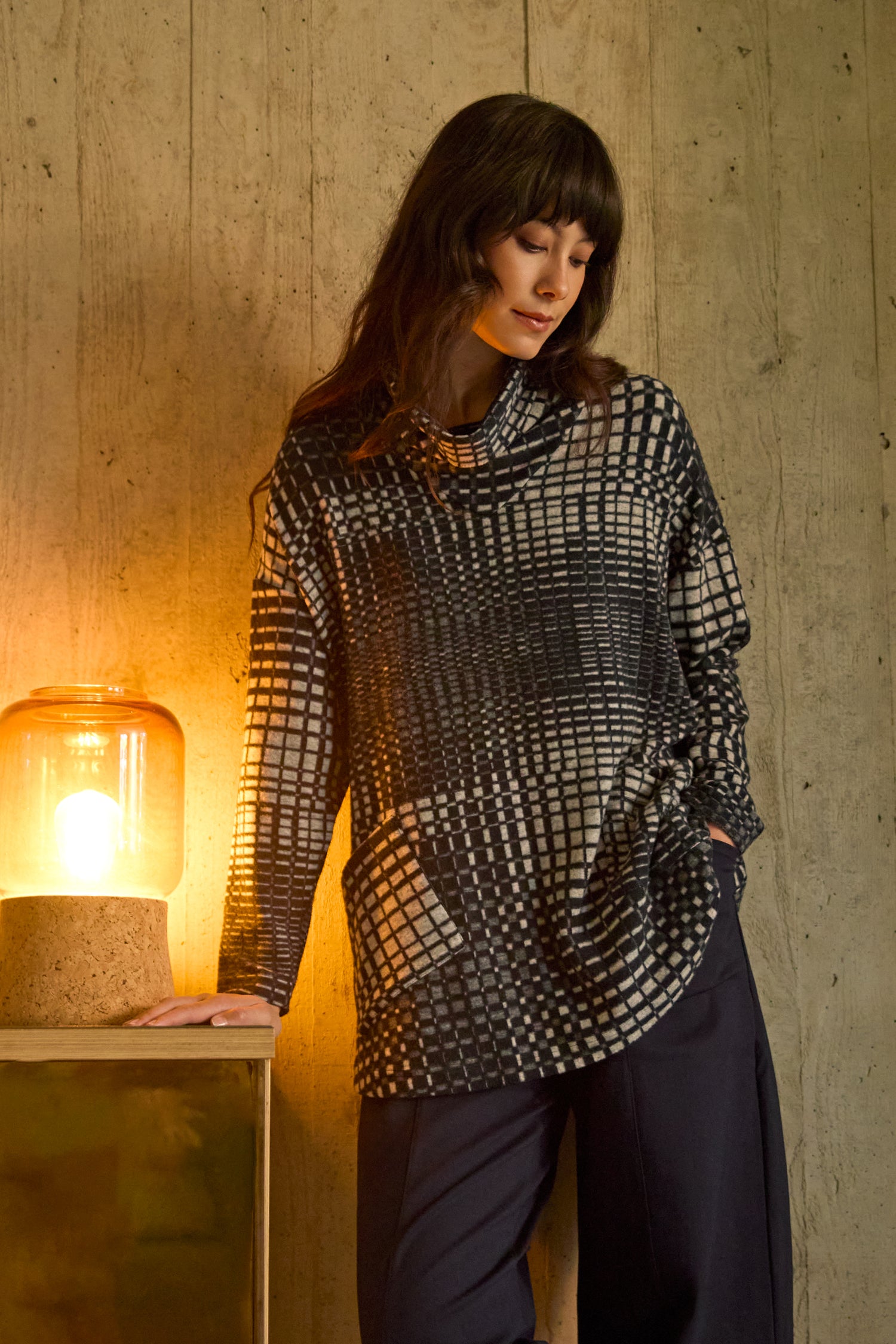A woman wears the Illusion Check Jersey Tunic with long sleeves and dark pants, standing by a lit lamp on a wooden table against a concrete wall.