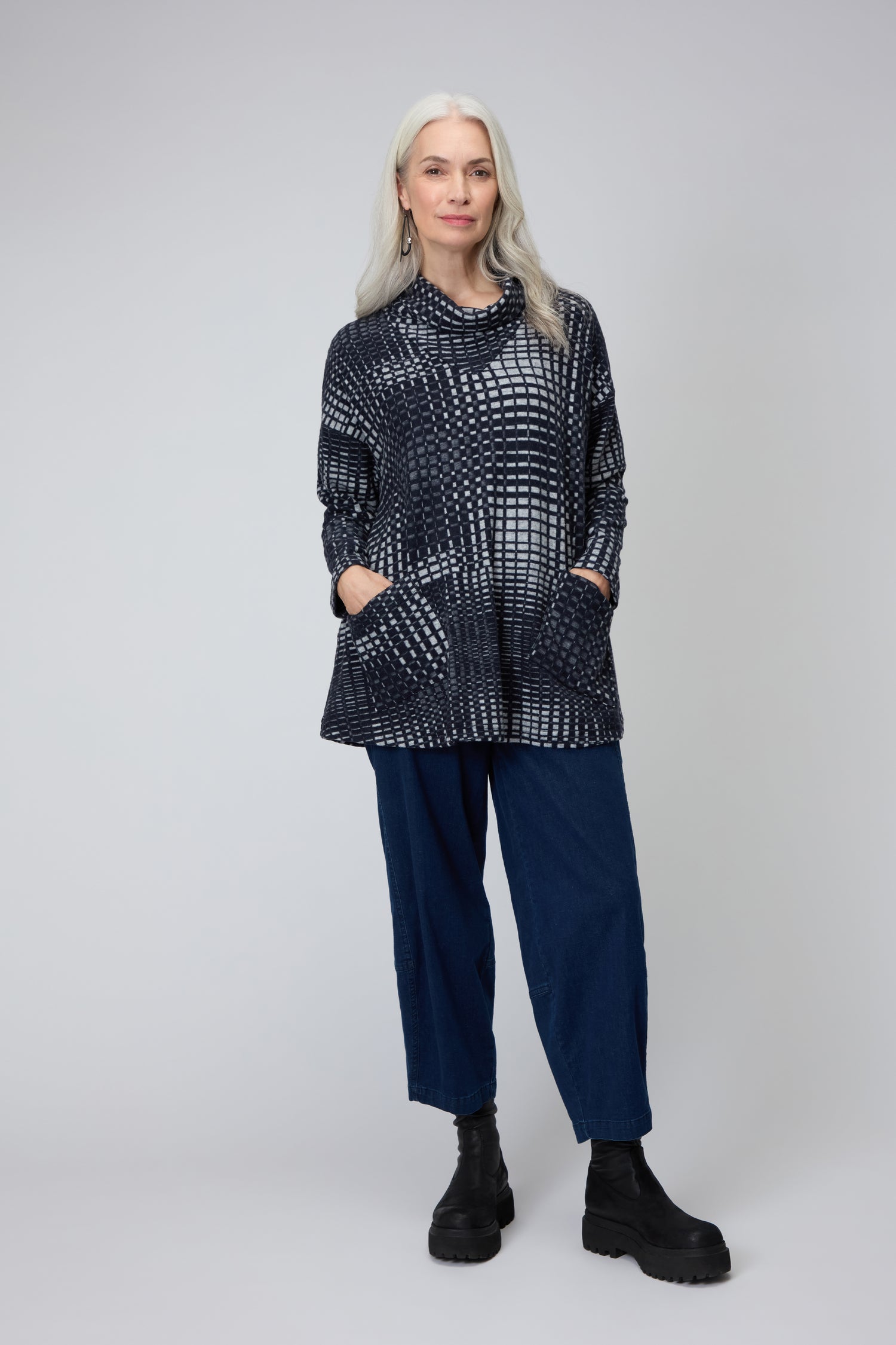 A woman with long gray hair wears the Illusion Check Jersey Tunic in black-and-white, paired with blue pants and black boots, standing against a plain gray background with her hands in her pockets.