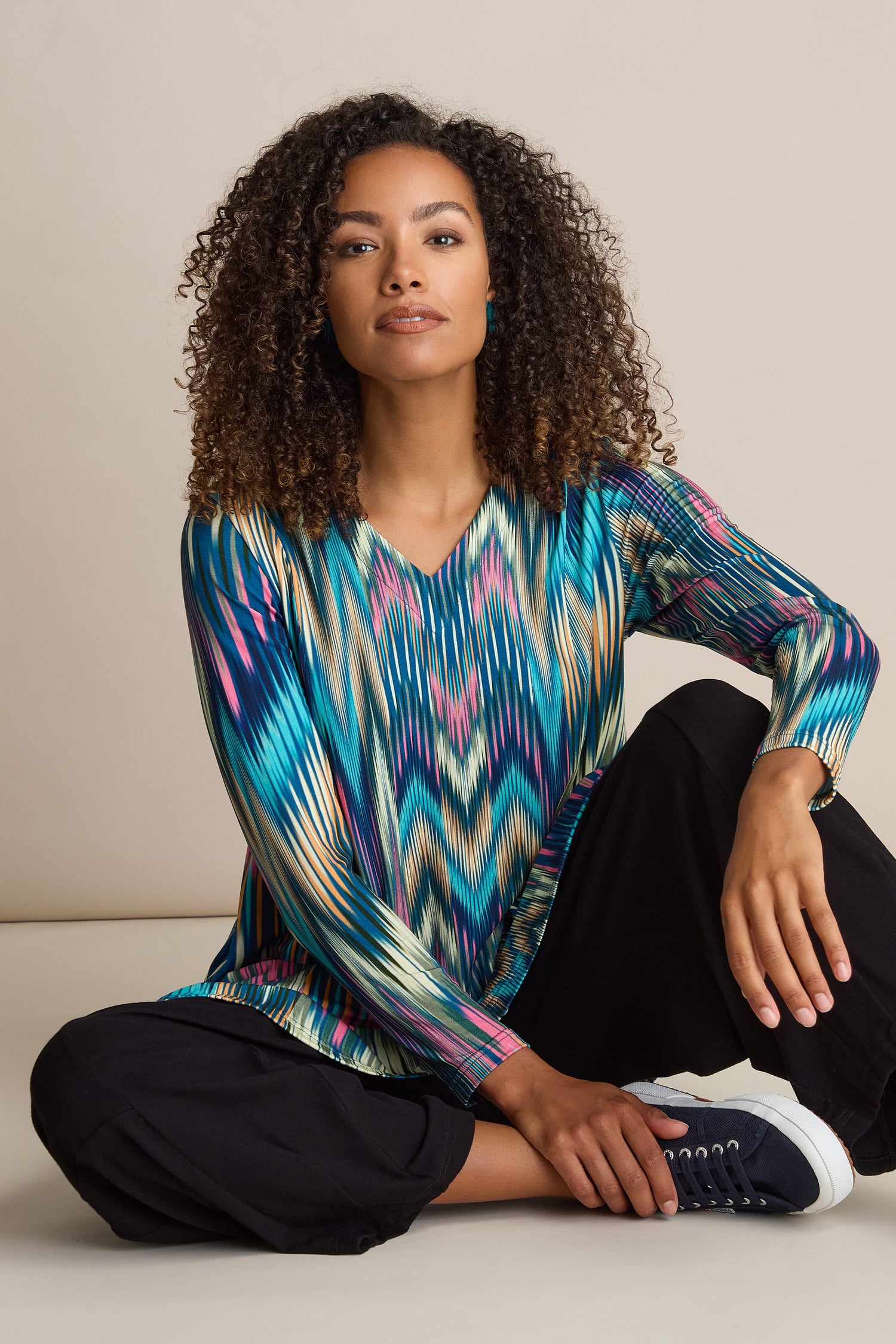 A person with curly hair sits on the floor, wearing an Autumnal Ikat Jersey Top and black pants, paired with dark sneakers—perfect for an Autumn wardrobe.