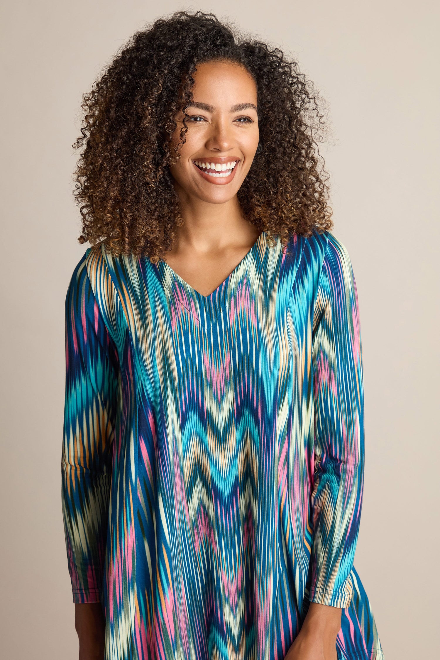 A person with curly hair smiles while looking slightly to the side against a plain background, showcasing the elegant Autumnal Ikat Jersey Top—an ideal addition to an autumn wardrobe.