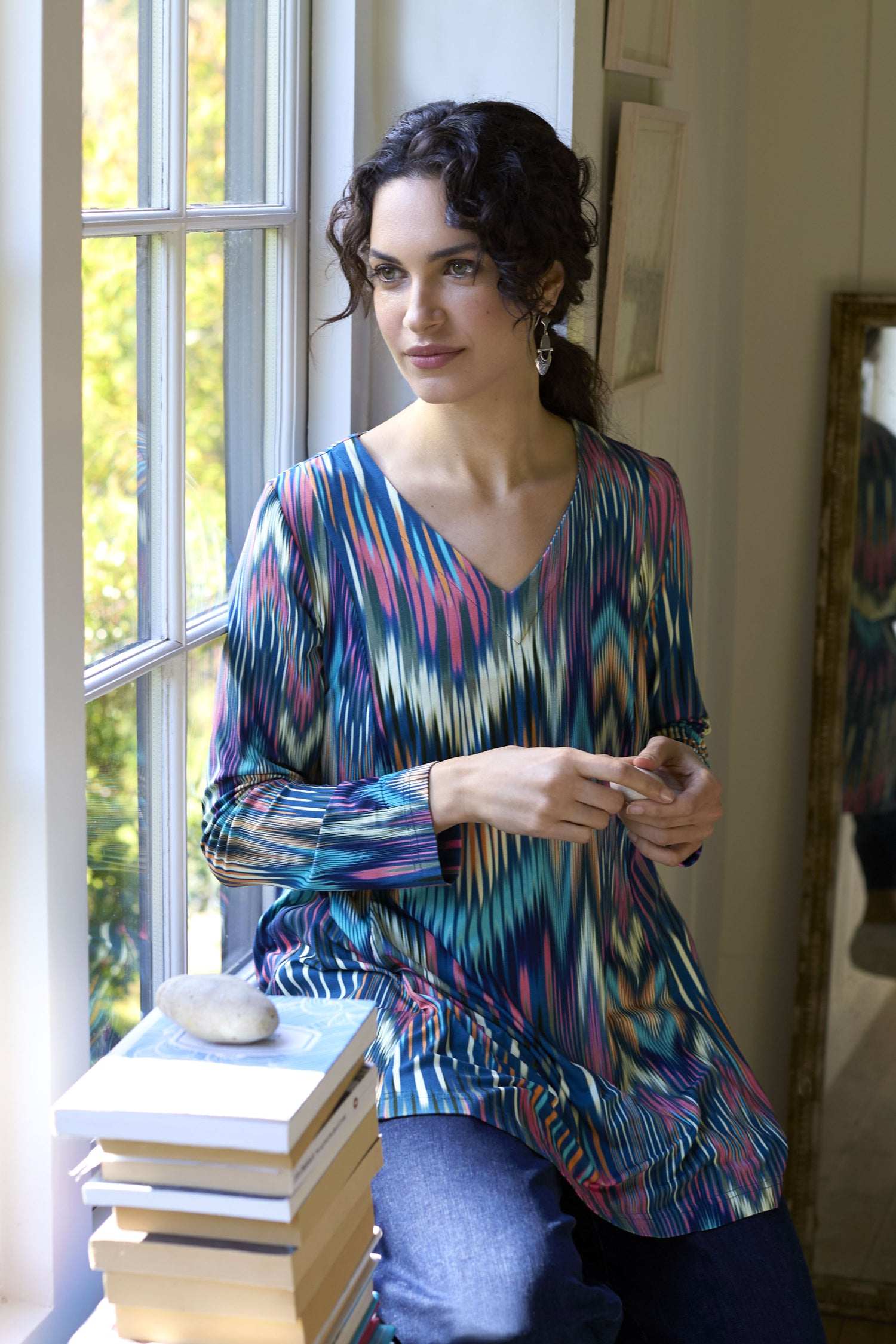 A person with dark curly hair and wearing an Autumnal Ikat Jersey Top sits by a window, holding an object. A stack of books and a stone are on the windowsill beside them, adding charm to their cozy autumn wardrobe.