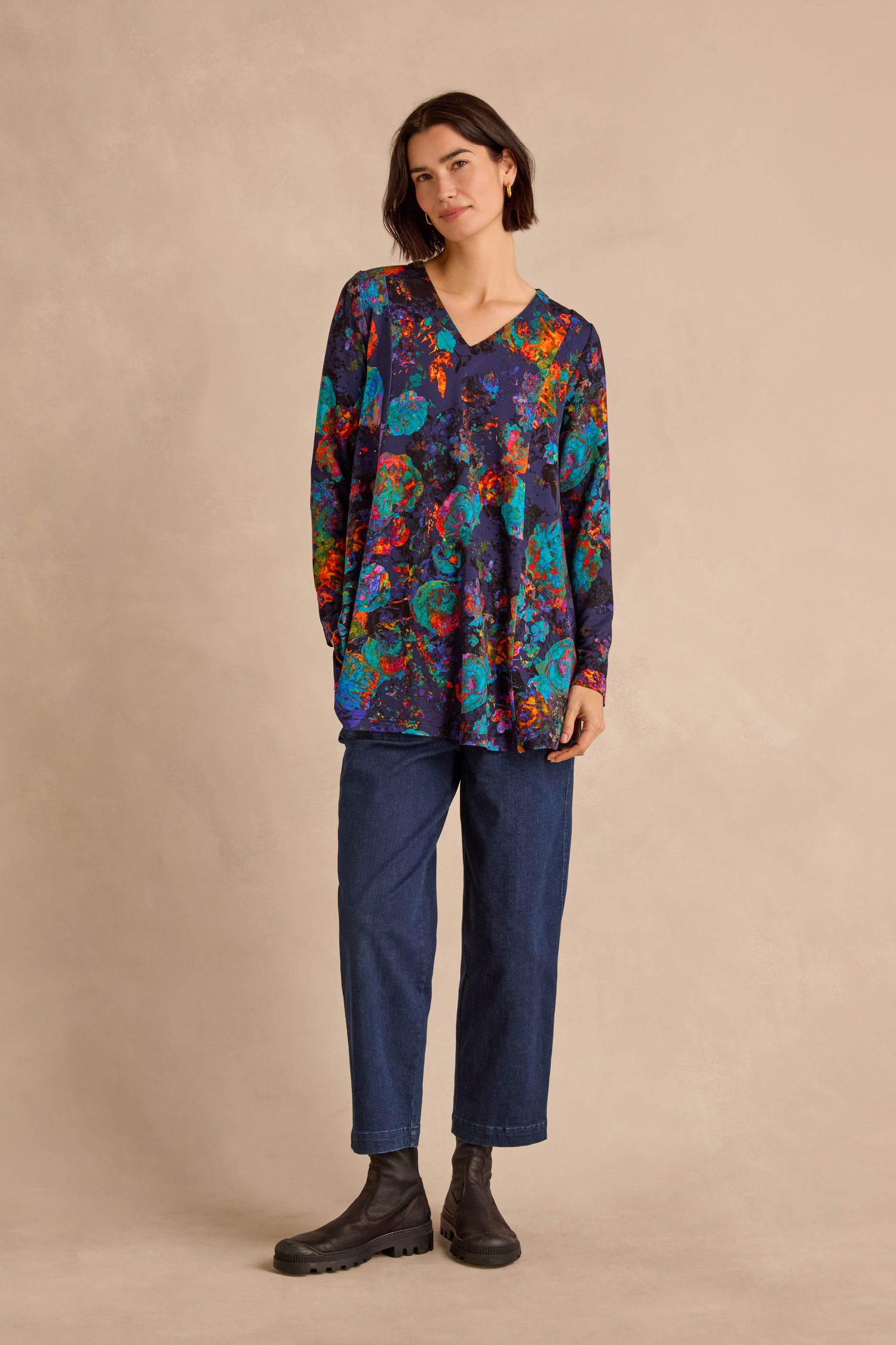 A woman stands against a beige background wearing the Midnight Floral Jersey Tunic—a loose, long-sleeved floral top—paired with blue jeans and black boots, her short dark hair framing a neutral expression.