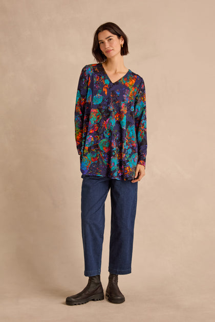A woman stands against a beige background wearing the Midnight Floral Jersey Tunic—a loose, long-sleeved floral top—paired with blue jeans and black boots, her short dark hair framing a neutral expression.