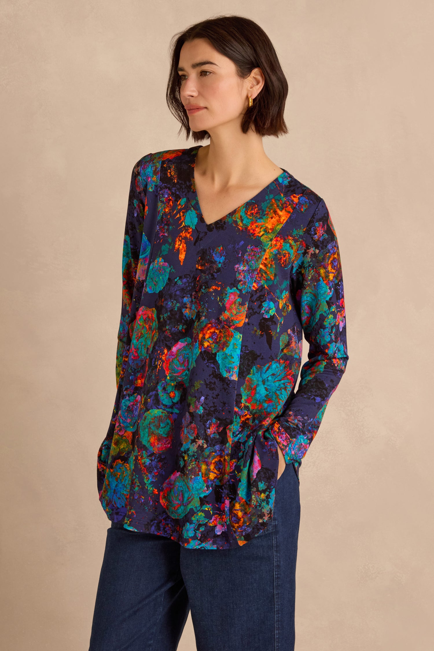A woman wearing the Midnight Floral Jersey Tunic—a long-sleeve, multicolored floral top—paired with blue jeans, stands with one hand in her pocket against a plain beige background.