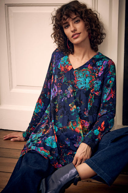 A woman wearing a Cosmic Peony Jersey Tunic with a vibrant, multicolored floral print on a dark blue background.