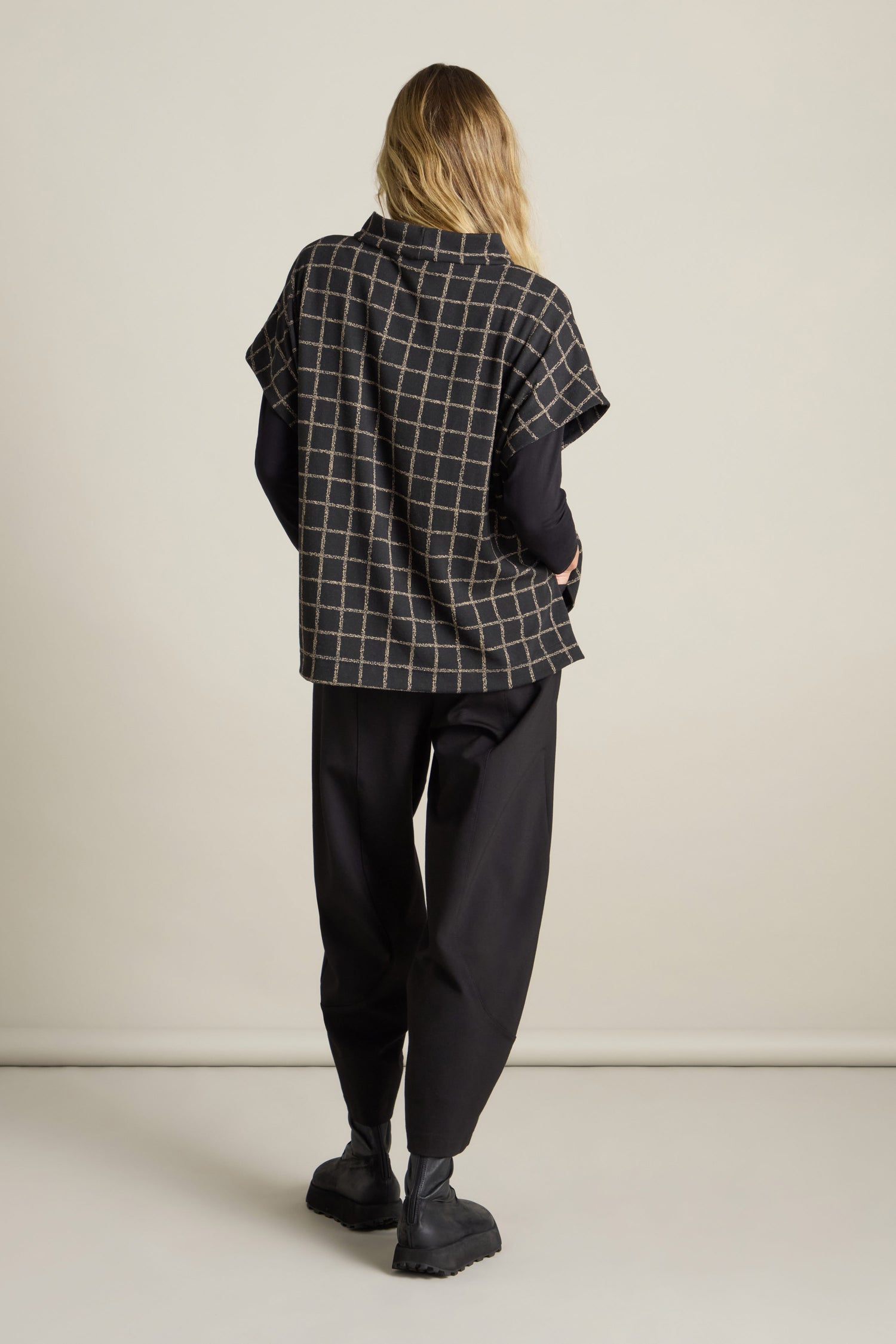 A person with long blonde hair stands facing away, wearing a black and brown Broken Check Jersey Tabard, paired with loose black pants and black shoes, against a plain, light-colored background. This stylish layering piece adds a modern edge to the overall look.