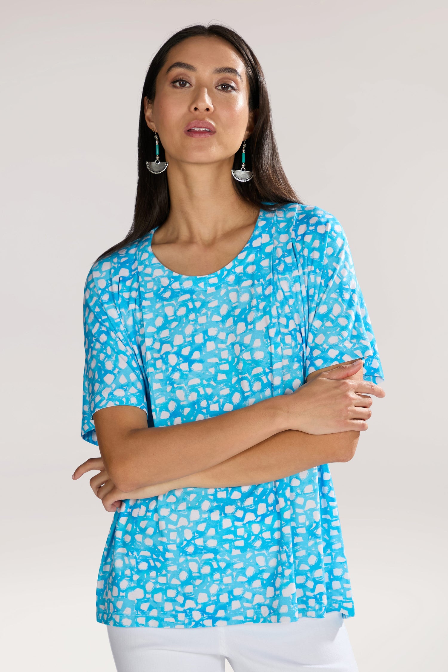 A person with long dark hair wearing a Pebble Jersey Print Relaxed Top featuring an abstract bubble motif and turquoise earrings, standing against a plain background.