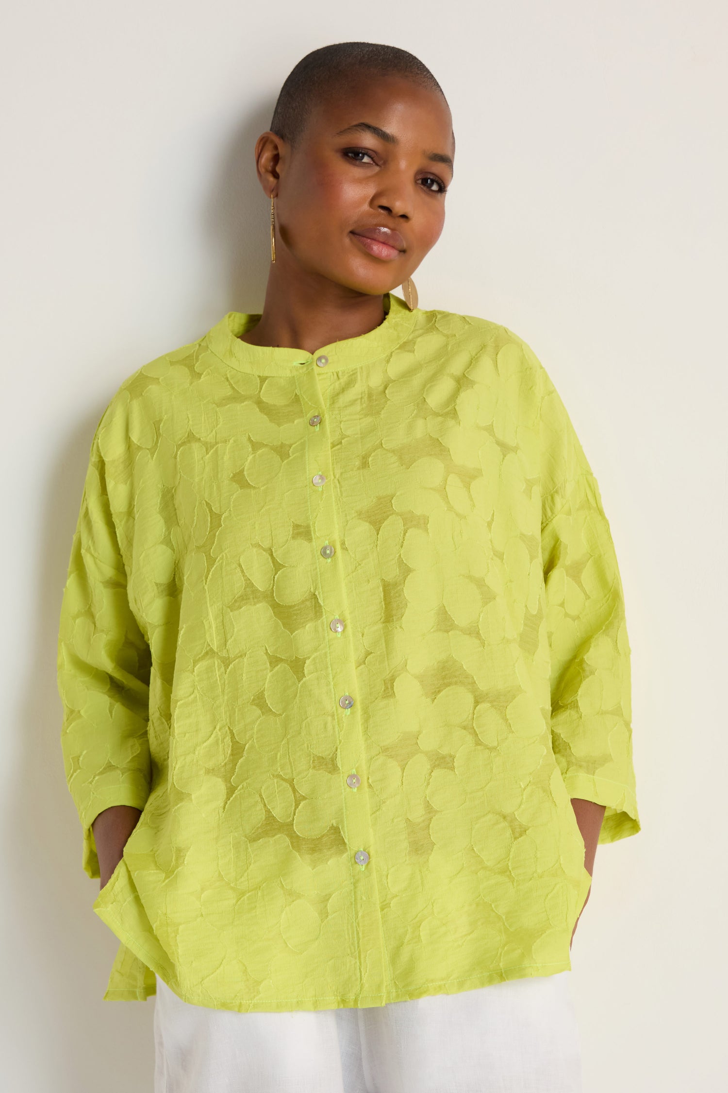 A person wears the Etched Floral Viscose Shirt—an oversized, light green top with three-quarter sleeves—paired with white pants and stands against a plain off-white background.