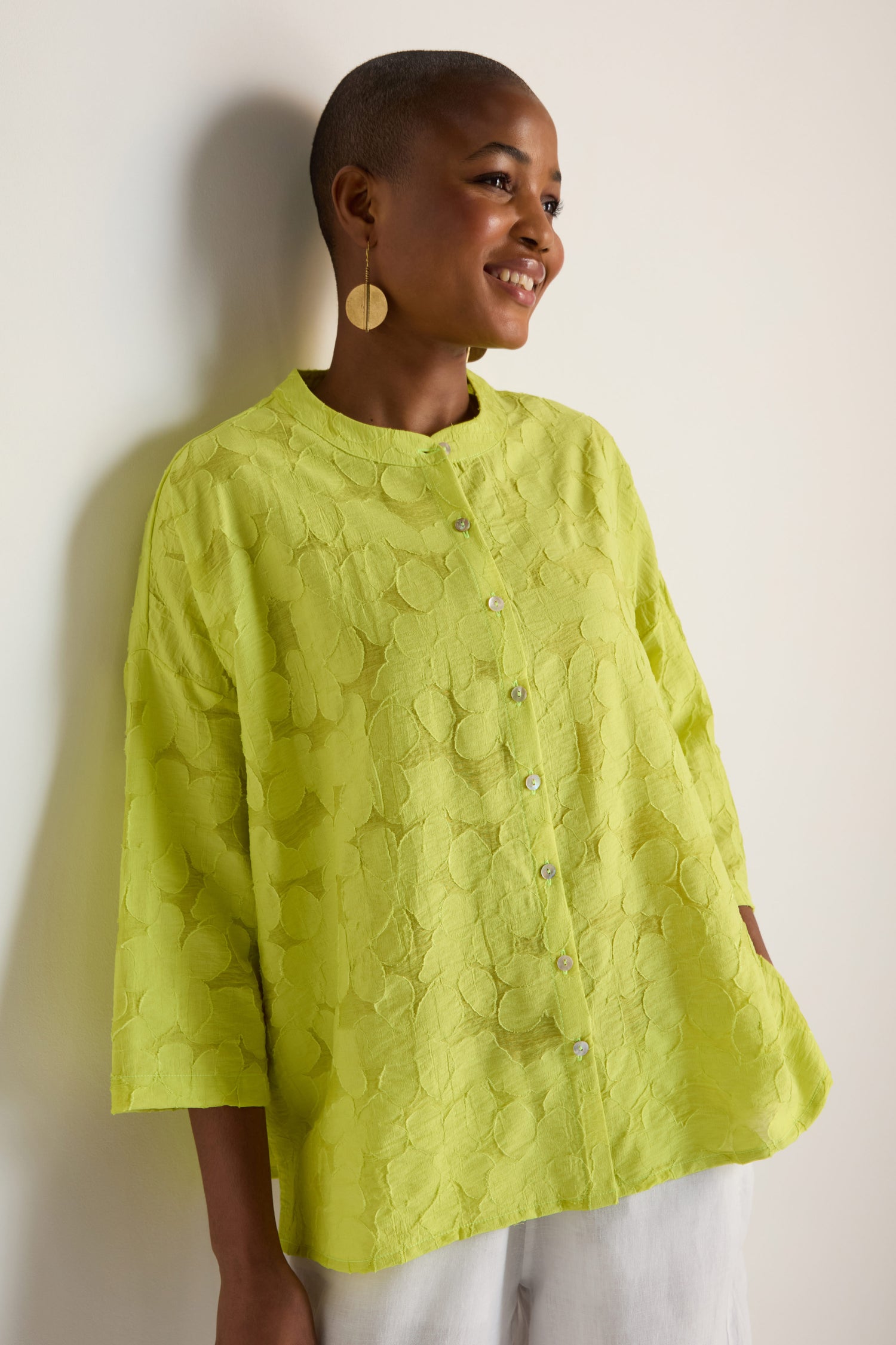 A person with short hair, smiling and wearing an oversized Etched Floral Viscose Shirt in textured yellow-green and large round earrings, leans against a white wall.
