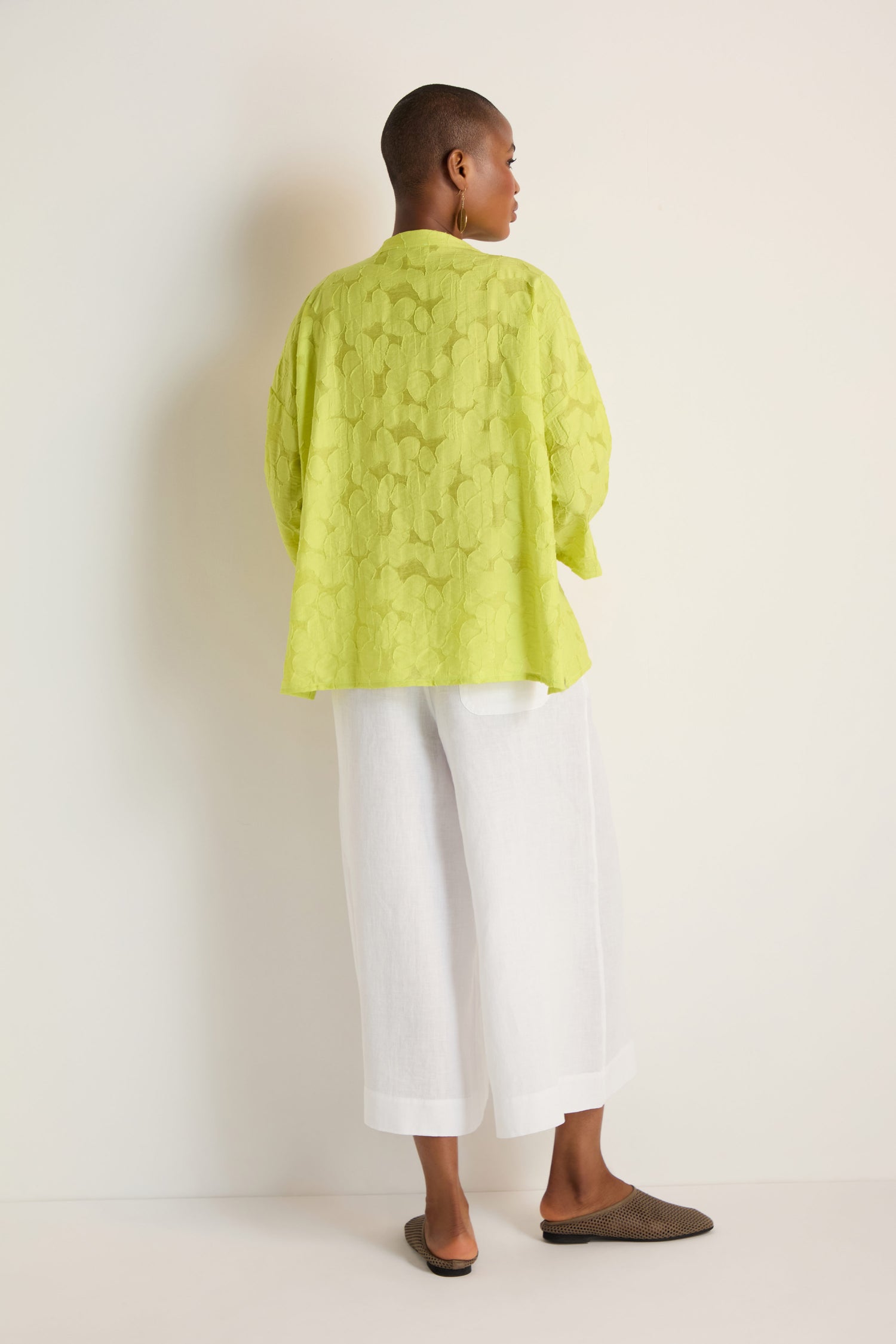 A person stands with their back to the camera, wearing the oversized Etched Floral Viscose Shirt in lime green, paired with wide white pants and brown slip-on shoes against a simple light background.