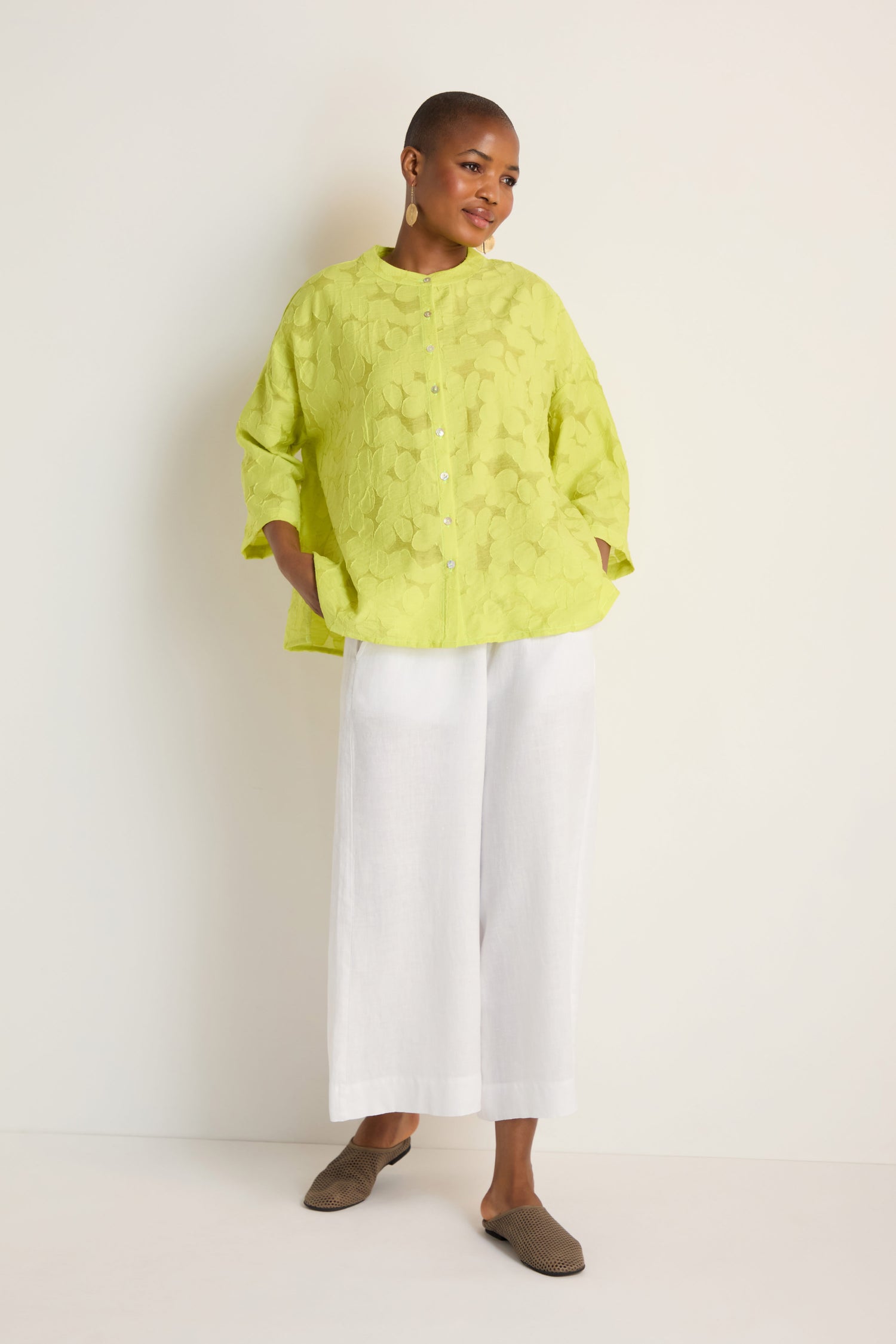 Person standing against a plain background wearing an oversized Etched Floral Viscose Shirt in yellow-green, paired with wide white pants, brown flats, and hoop earrings.