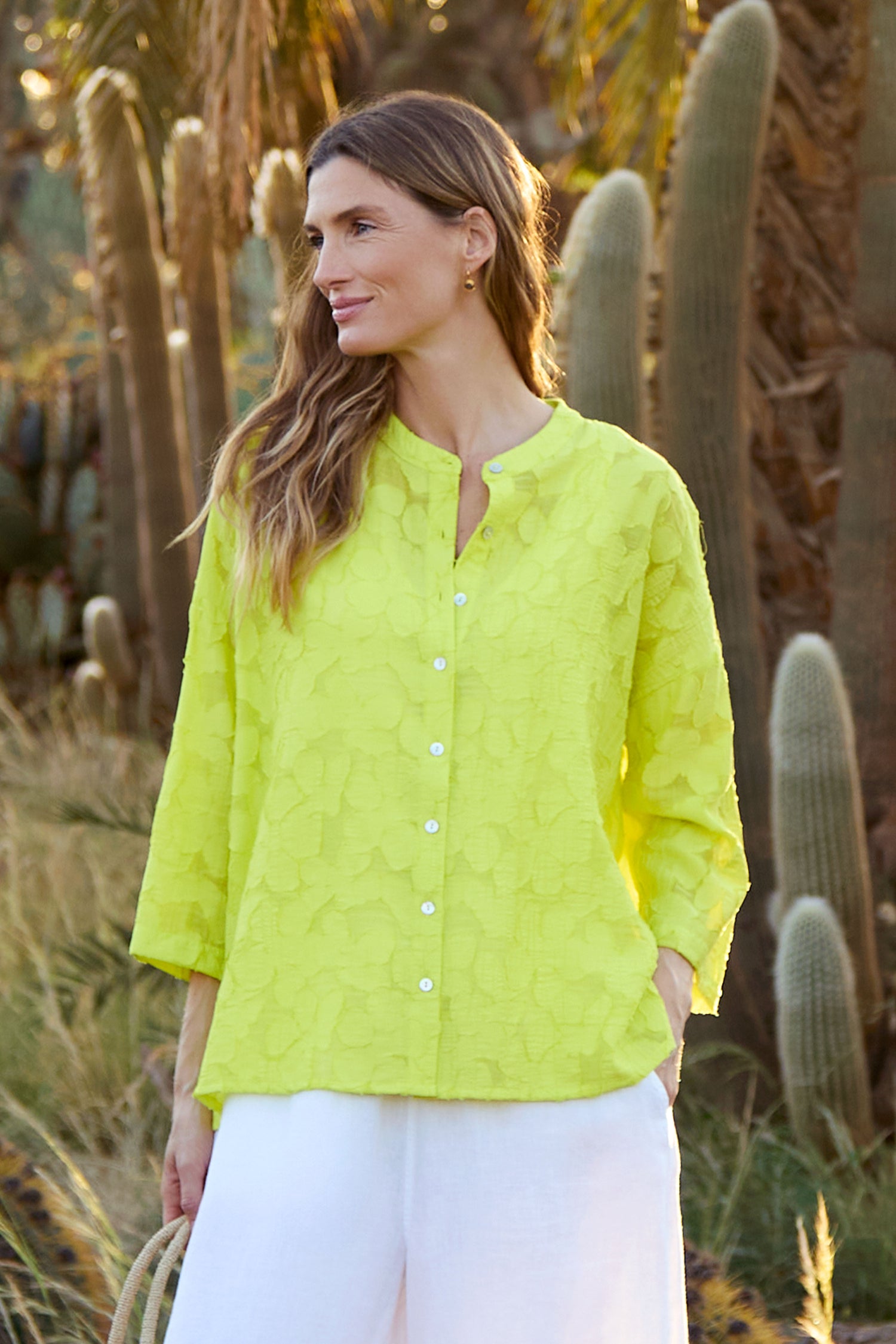 A woman stands outdoors among tall cacti and grass, looking to the side in an oversized Etched Floral Viscose Shirt paired with white pants.