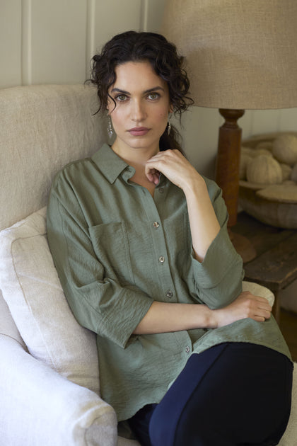 A person with curly hair sits on a beige couch wearing a relaxed chic, green Textured Viscose Boxy Shirt and dark pants, with one hand touching their face.