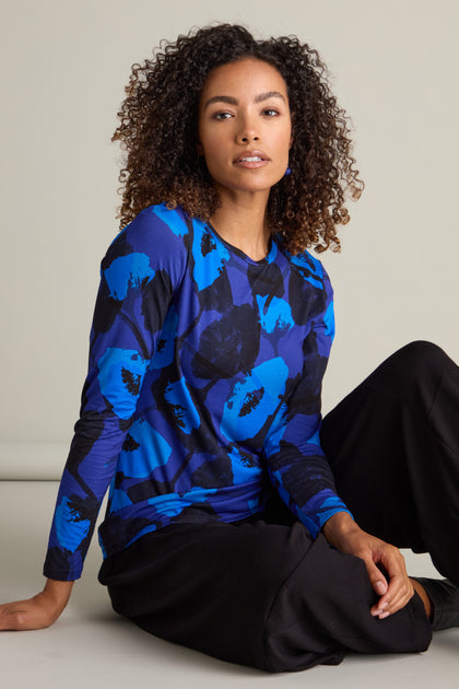 A woman with curly hair, seated on the floor, is wearing a blue Flowering Poppies Jersey Top and black slim-fit pants.