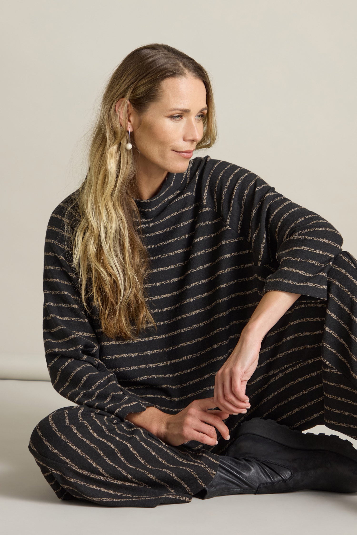 A person with long, wavy hair sits on the floor, wearing a black Broken Stripe Jersey Top detailed with gold horizontal stripes and a mock neck, gazing to the side.