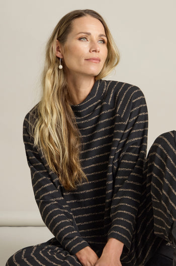 A woman with long blonde hair, adorned in a black and gold Broken Stripe Jersey Top, sits while gazing slightly to the right.