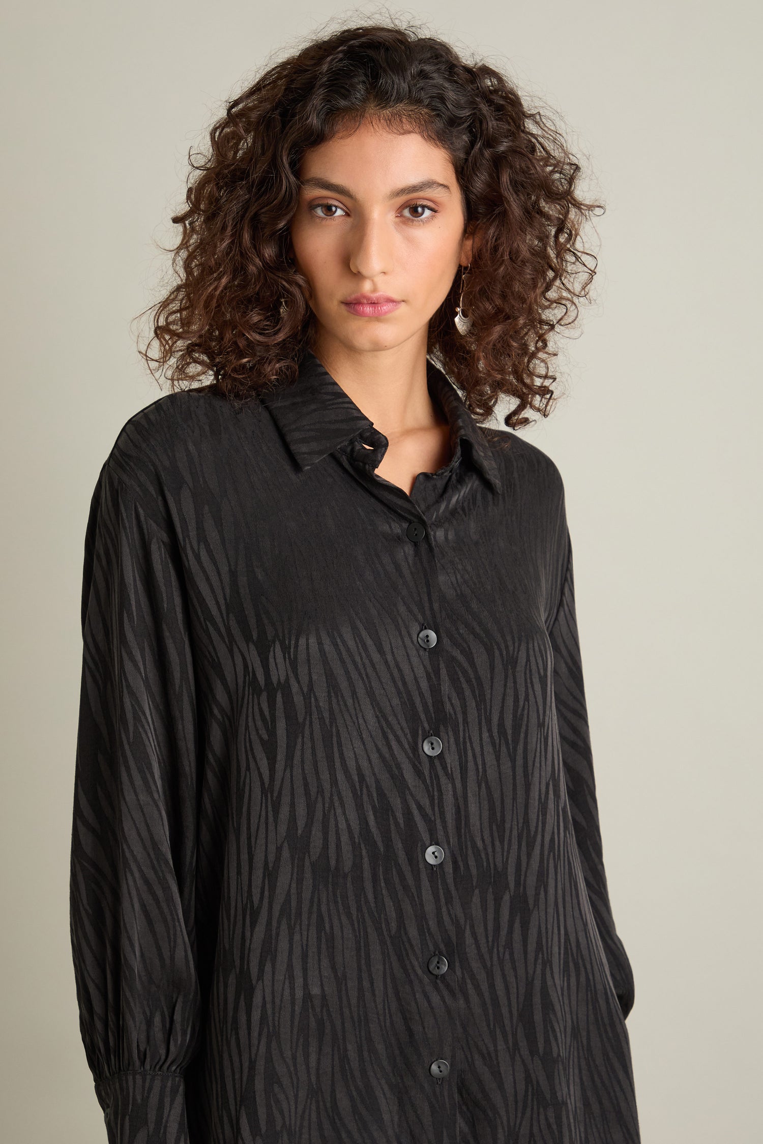 Portrait of a person with curly hair wearing the Abstract Lines Jacquard Long Shirt, standing against a neutral background. The black, patterned button-down shirt exhibits a relaxed fit and silky-feel fabric.