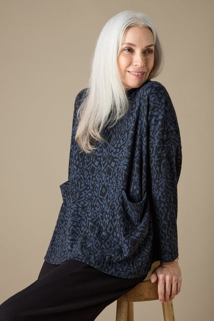 A person with long white hair is sitting on a stool, wearing the Ikat Soft Jersey Relaxed Top in dark blue abstract print along with black pants, facing slightly left and smiling.