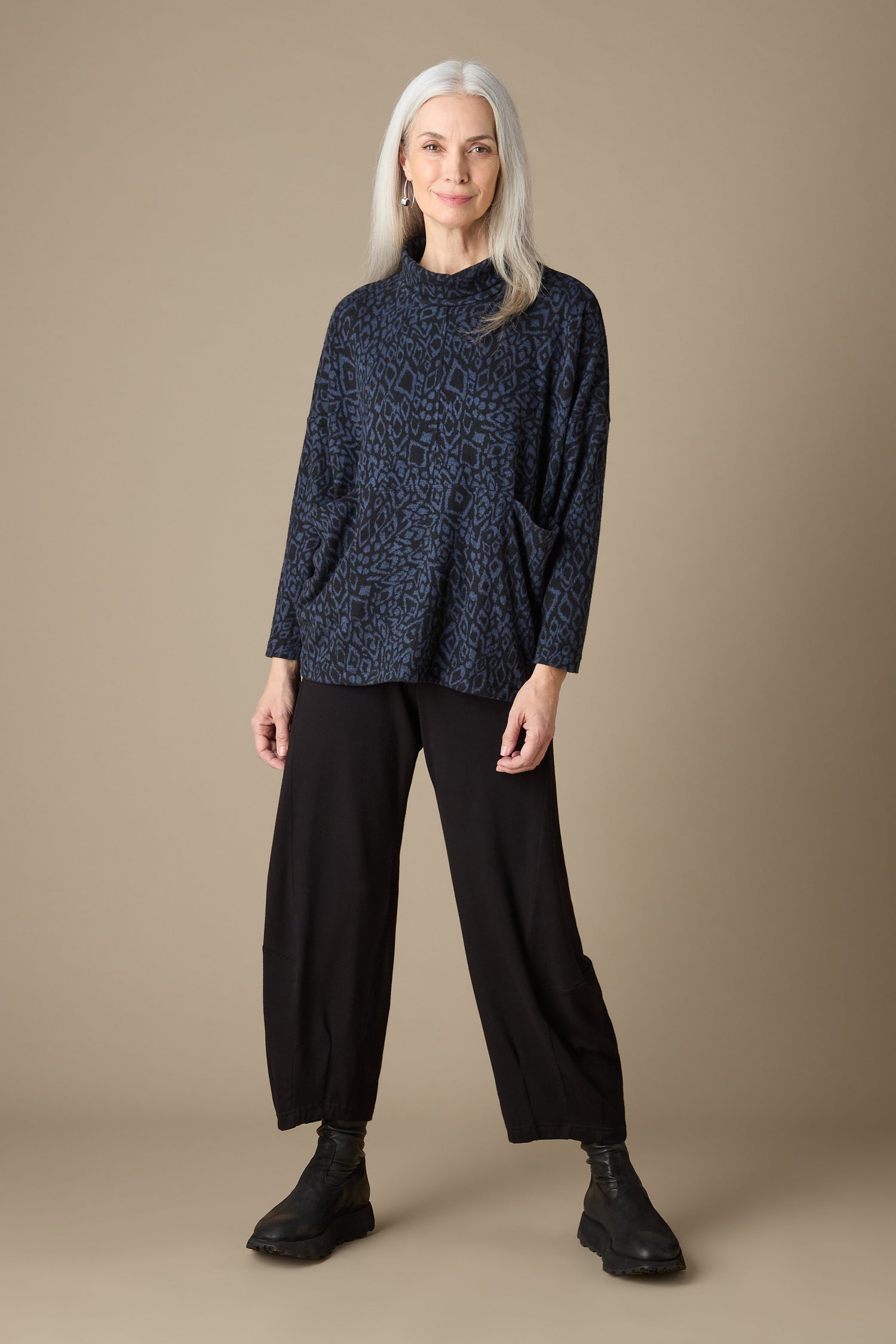 An older woman with long gray hair stands confidently, showcasing the Ikat Soft Jersey Relaxed Top, which features an abstract print. She pairs it elegantly with black pants and shoes against a plain beige background, exuding quiet elegance and comfort.