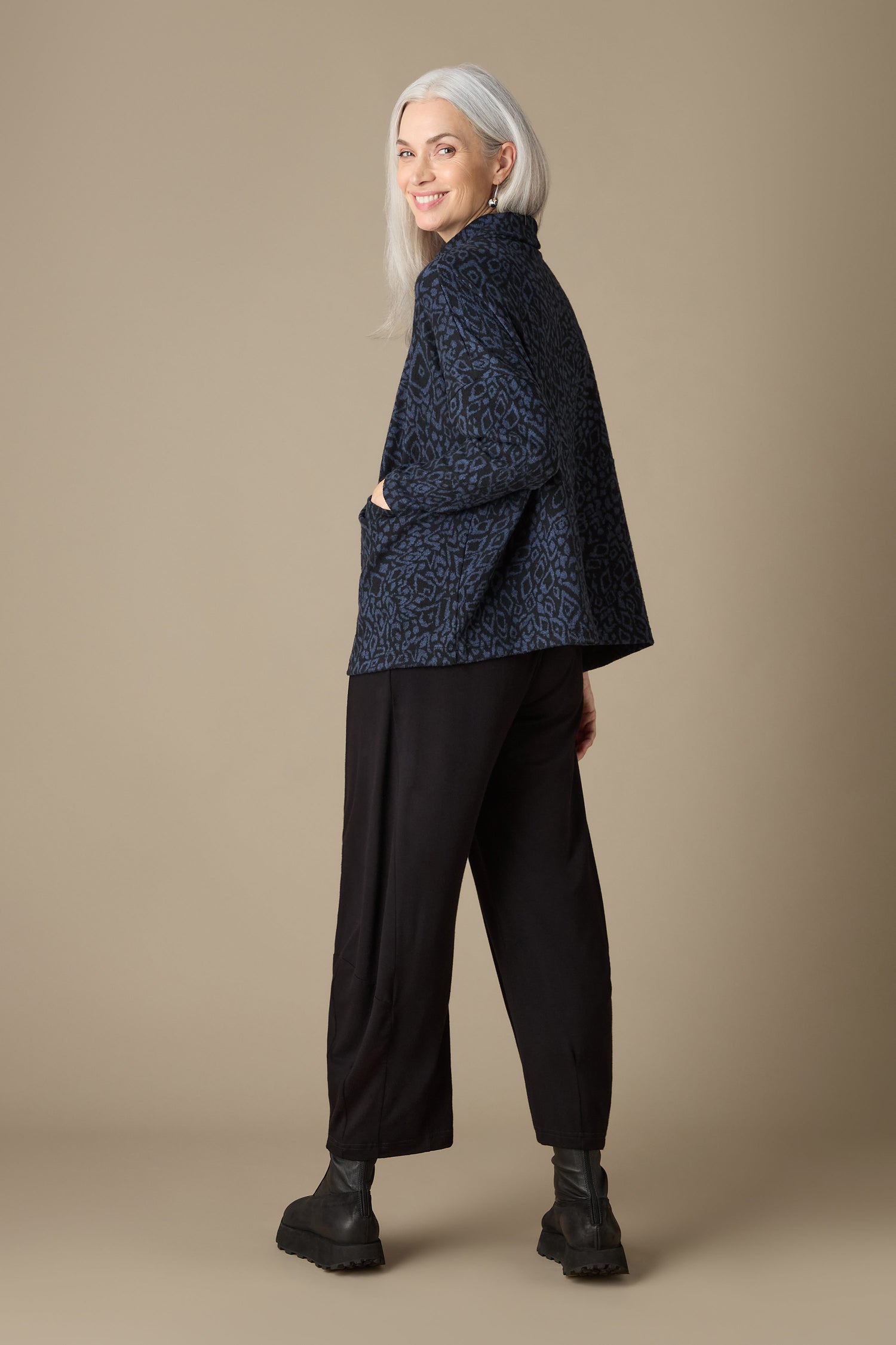 A woman with gray hair smiles while wearing the Ikat Soft Jersey Relaxed Top, paired with black pants and boots, as she stands against a plain beige background.