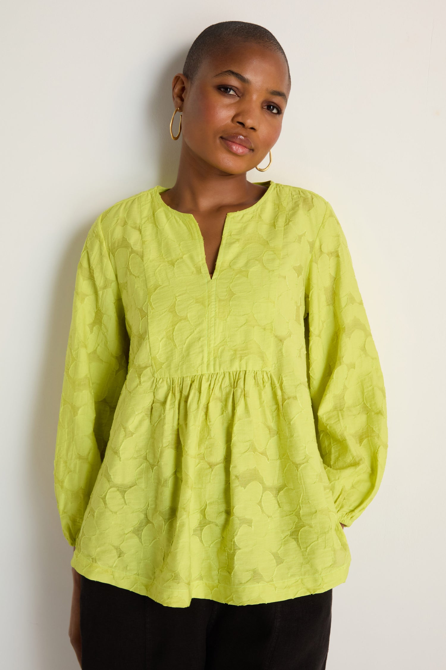A person with short hair and gold hoop earrings wears the Etched Floral Viscose Top in a yellow-green woven floral pattern, standing against a plain white wall.