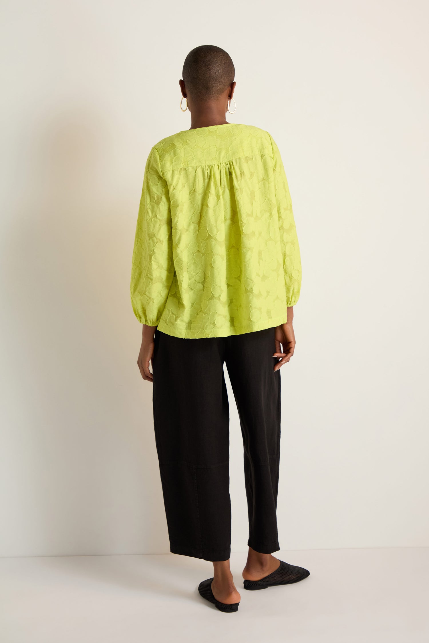 A person stands against a white wall, seen from the back, wearing a bright yellow Etched Floral Viscose Top, loose black pants, black shoes, and hoop earrings.