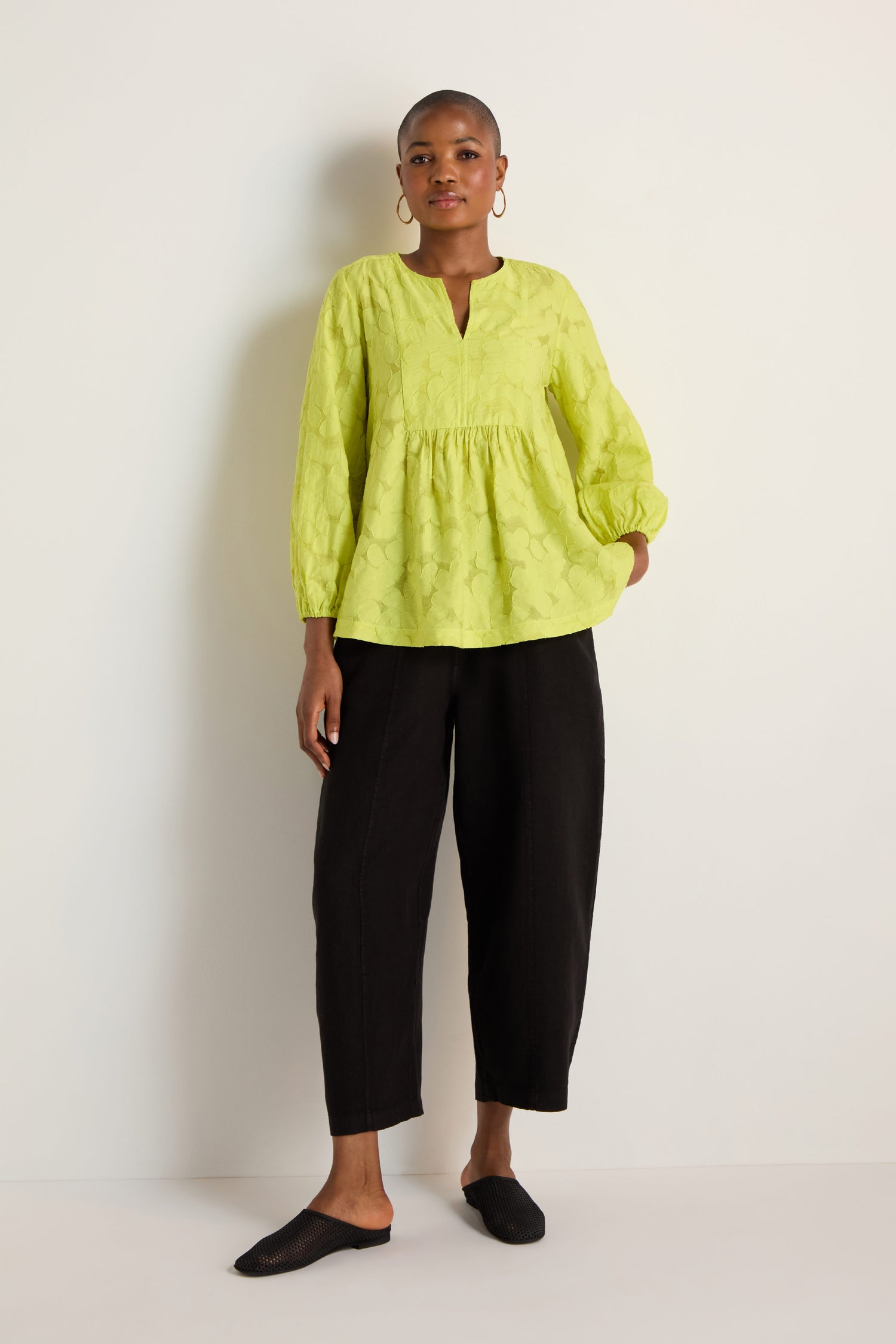 A person stands against a plain wall wearing the Etched Floral Viscose Top in light green, paired with black cropped pants and black slip-on shoes.