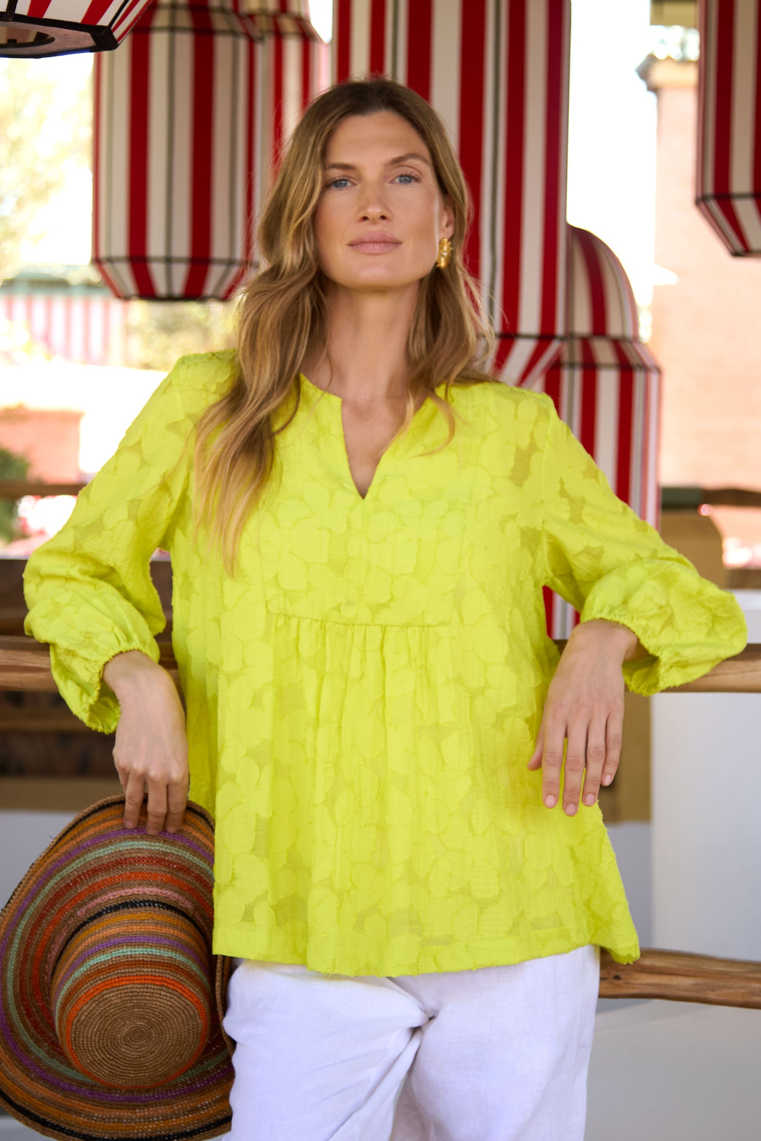 A woman indoors wears the Etched Floral Viscose Top in bright yellow and white pants, holding a multicolored hat, with red and white striped decor in the background.