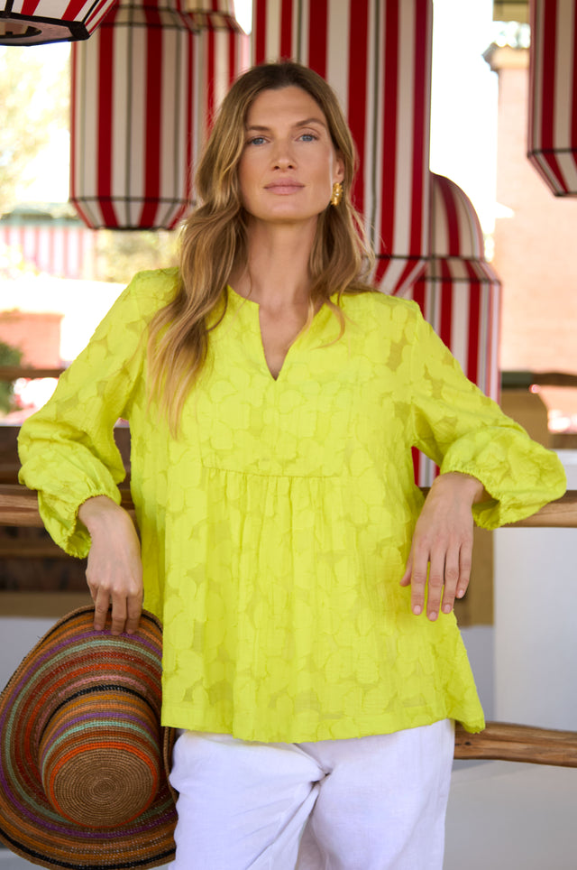 A woman indoors wears the Etched Floral Viscose Top in bright yellow and white pants, holding a multicolored hat, with red and white striped decor in the background.