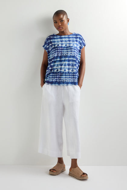 Wearing a blue Tie Dye Jersey Cold Shoulder Top with an oversized fit, the person pairs it with white wide-legged pants and tan sandals, standing against a crisp white background.