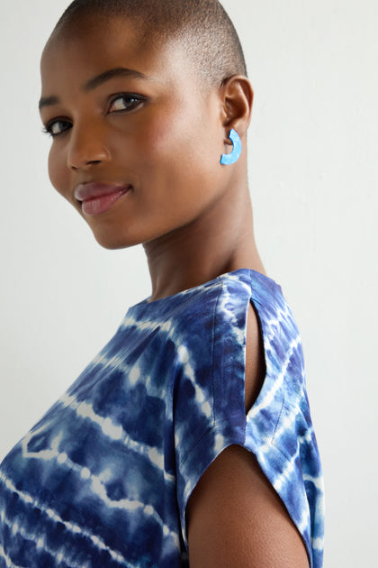 A person with short hair smiles at the camera, wearing a Tie Dye Jersey Cold Shoulder Top in blue shades, paired with matching blue earrings against a plain backdrop.