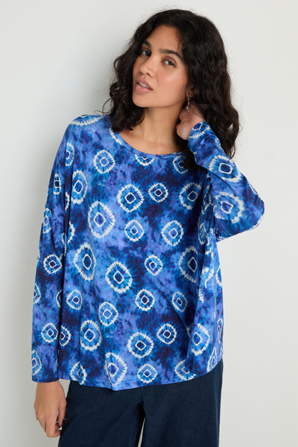 Woman wearing the Tie Dye Circles Jersey Boxy Top, a long-sleeve blue viscose jersey with a boxy fit, paired with dark pants and standing against a light-colored background.