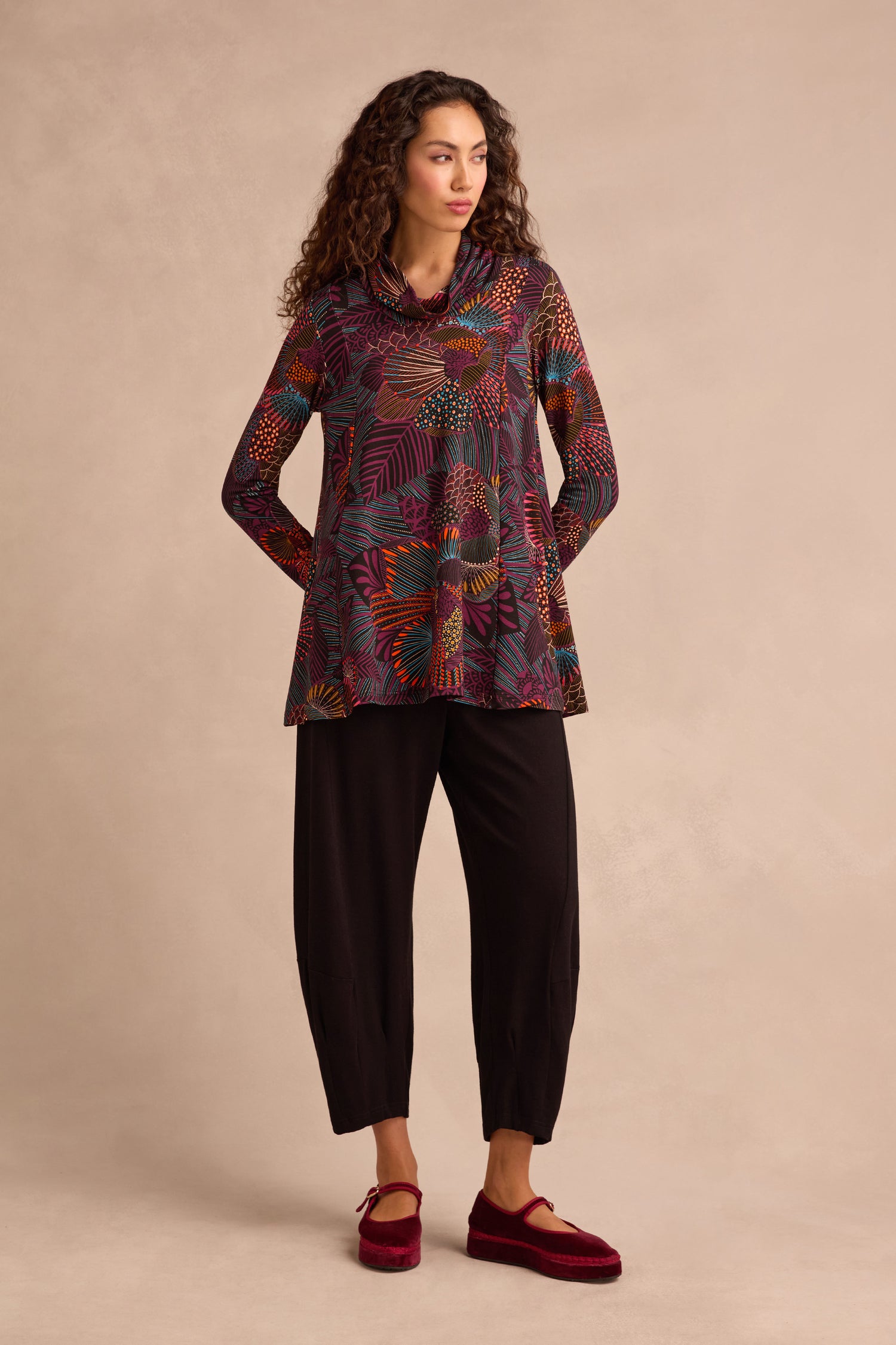 A person stands against a plain beige background wearing the Abstract Linear Floral Jersey Tunic featuring a multicolored, floral print and fit-and-flare silhouette, paired with black cropped pants and red shoes.