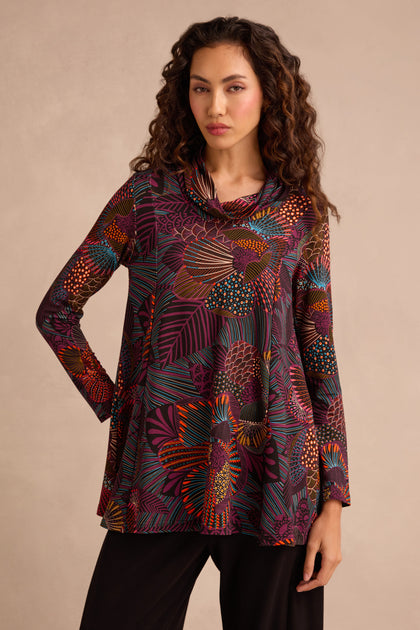 A woman is wearing the Abstract Linear Floral Jersey Tunic, featuring a long-sleeve, fit and flare silhouette with abstract floral and leaf patterns in red, orange, purple, and yellow shades. She has paired it with black pants and stands against a neutral background.