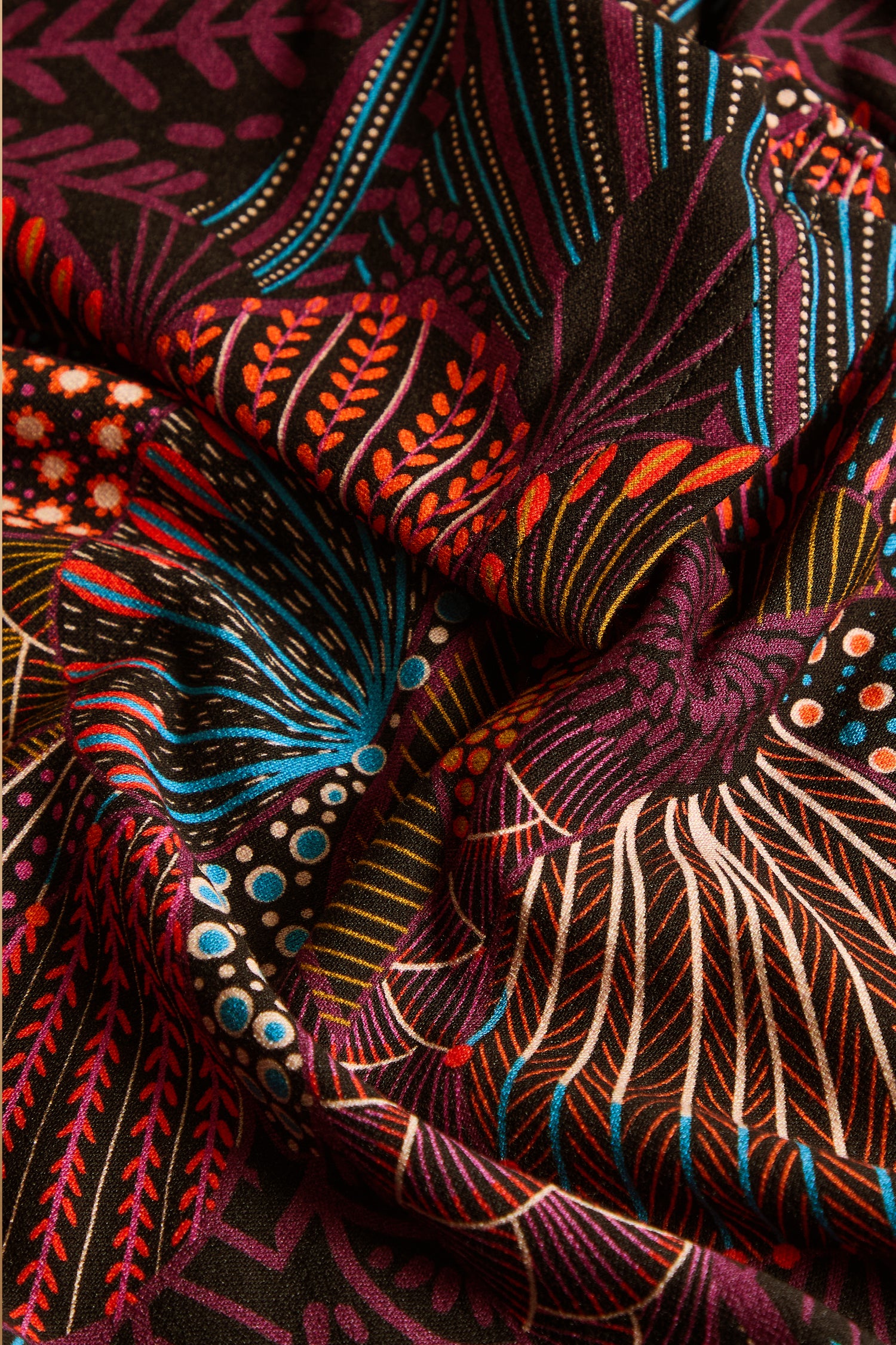 Close-up of colorful, patterned fabric with abstract floral and leaf designs in red, blue, orange, yellow, and white on a dark background. This fabric—used in the Abstract Linear Floral Jersey Tunic—features gentle folds and slight wrinkles that would beautifully complement a fit and flare silhouette.