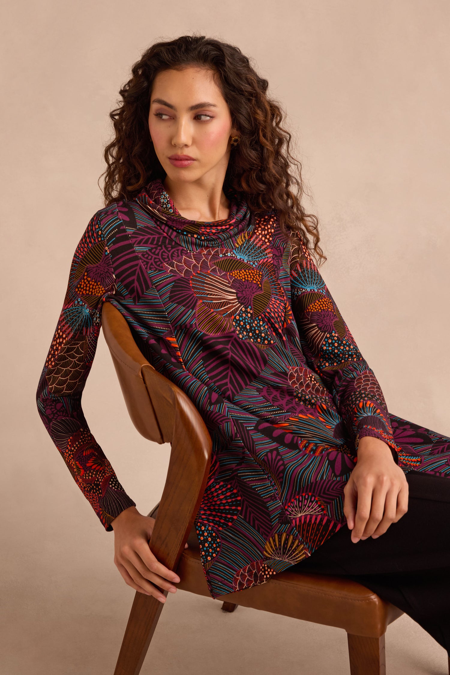A woman seated on a wooden chair, wearing the Abstract Linear Floral Jersey Tunic—a long-sleeved, multicolored floral top with a fit and flare silhouette—paired with black pants, looking to the side against a neutral background.