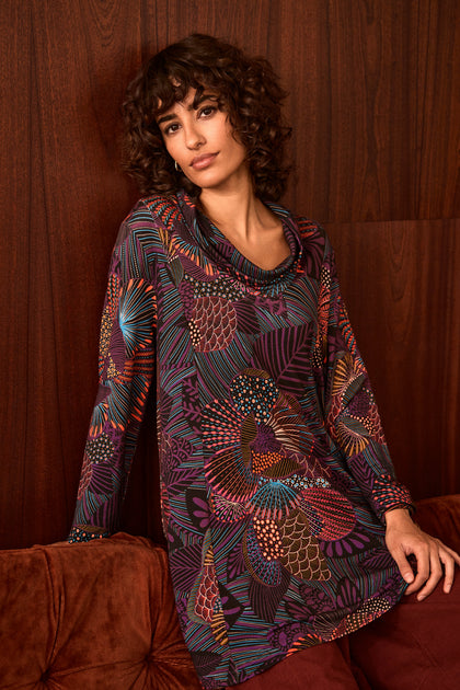 A woman with curly hair sits on a brown couch in front of a dark wood-paneled wall, wearing the Abstract Linear Floral Jersey Tunic—a colorful long-sleeve top with a draped cowl neck—paired with burgundy pants.