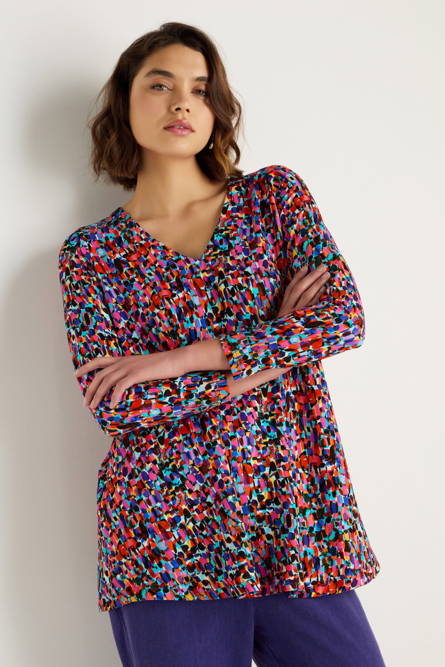 A woman stands against a plain wall, arms crossed, wearing the Painted Spots Jersey Tunic with long sleeves and purple pants.
