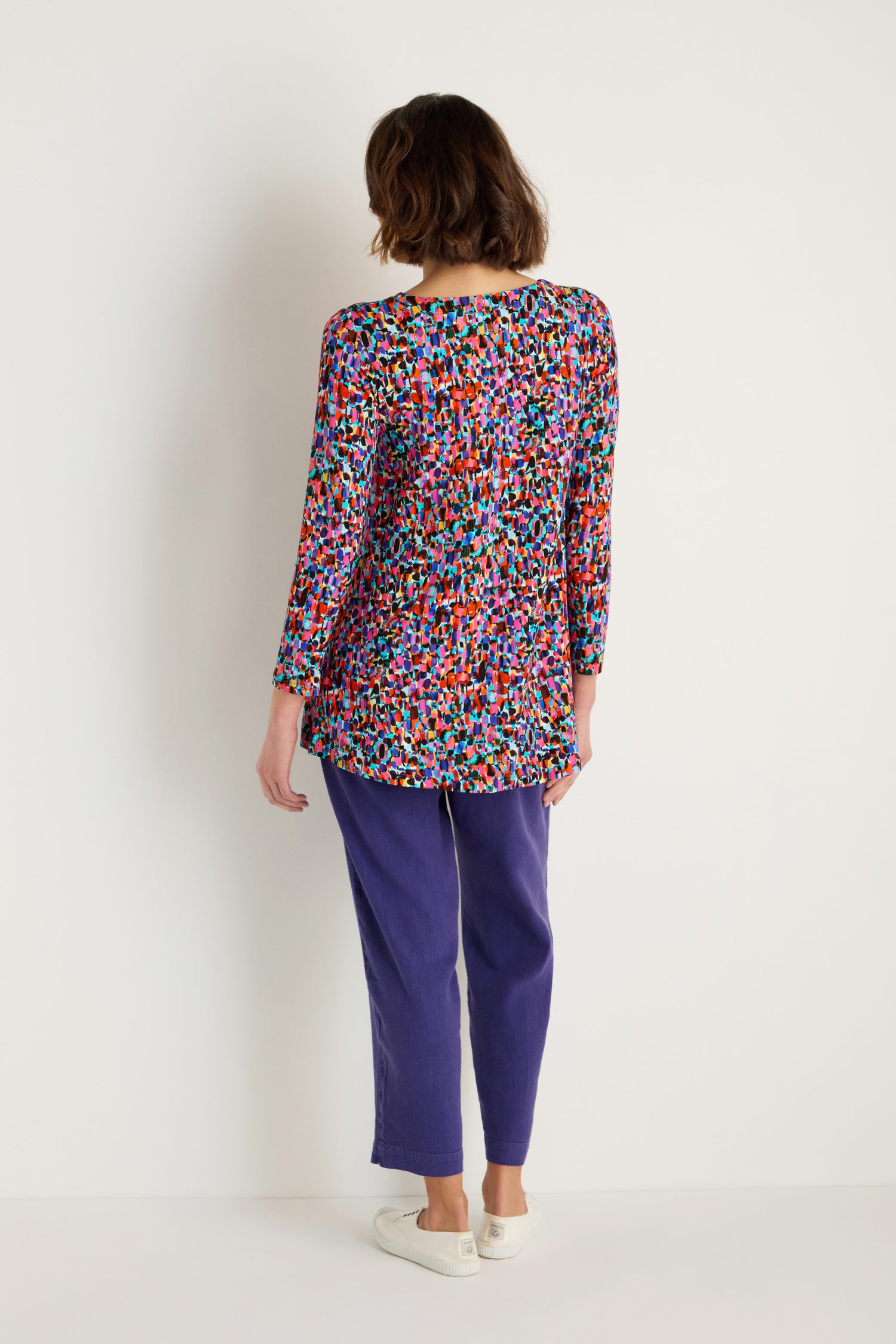 A person stands facing away, wearing the Painted Spots Jersey Tunic with purple pants and white slip-on shoes against a plain light background.
