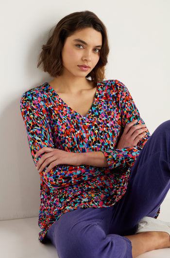 Woman sitting with arms crossed, wearing the Painted Spots Jersey Tunic and purple pants, against a plain light-colored background.