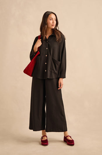 A woman stands facing left, wearing the Mini Crinkle Viscose Boxy Shirt in black paired with wide-leg pants, red velvet shoes, and a red shoulder bag, set against a neutral beige background.