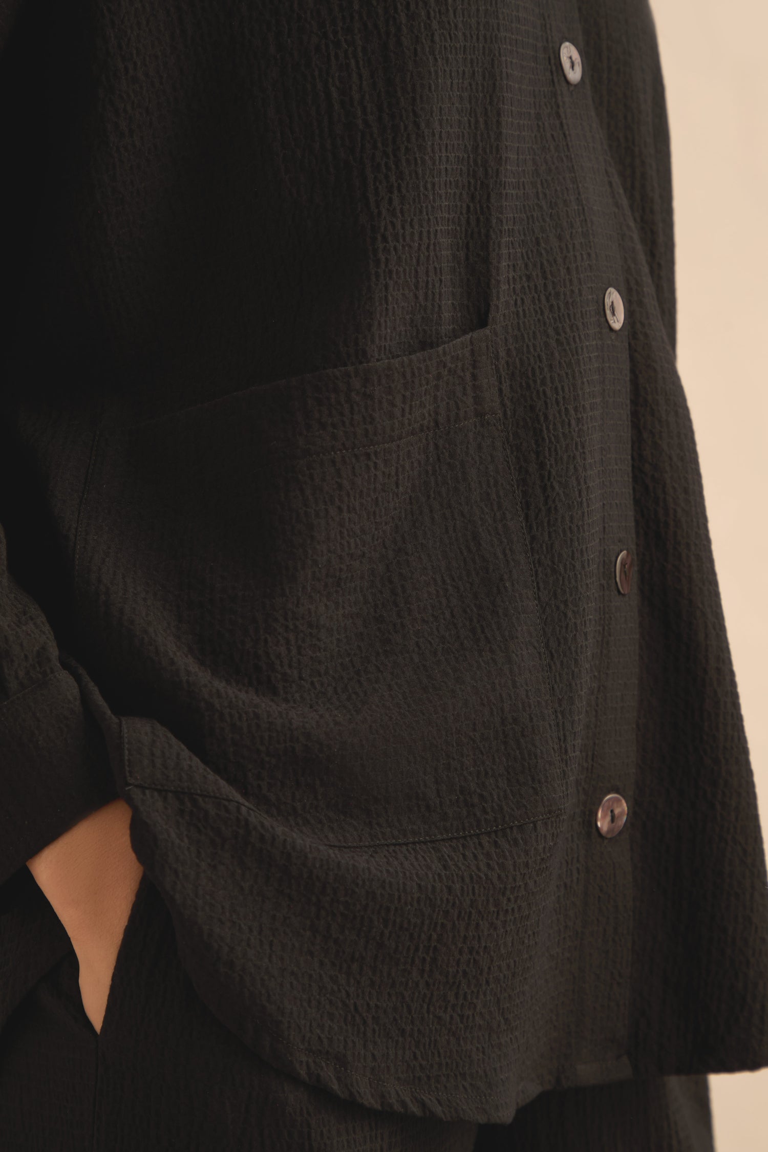 Close-up of a person wearing the Mini Crinkle Viscose Boxy Shirt in textured black with a front pocket, their hand tucked into their pants pocket.