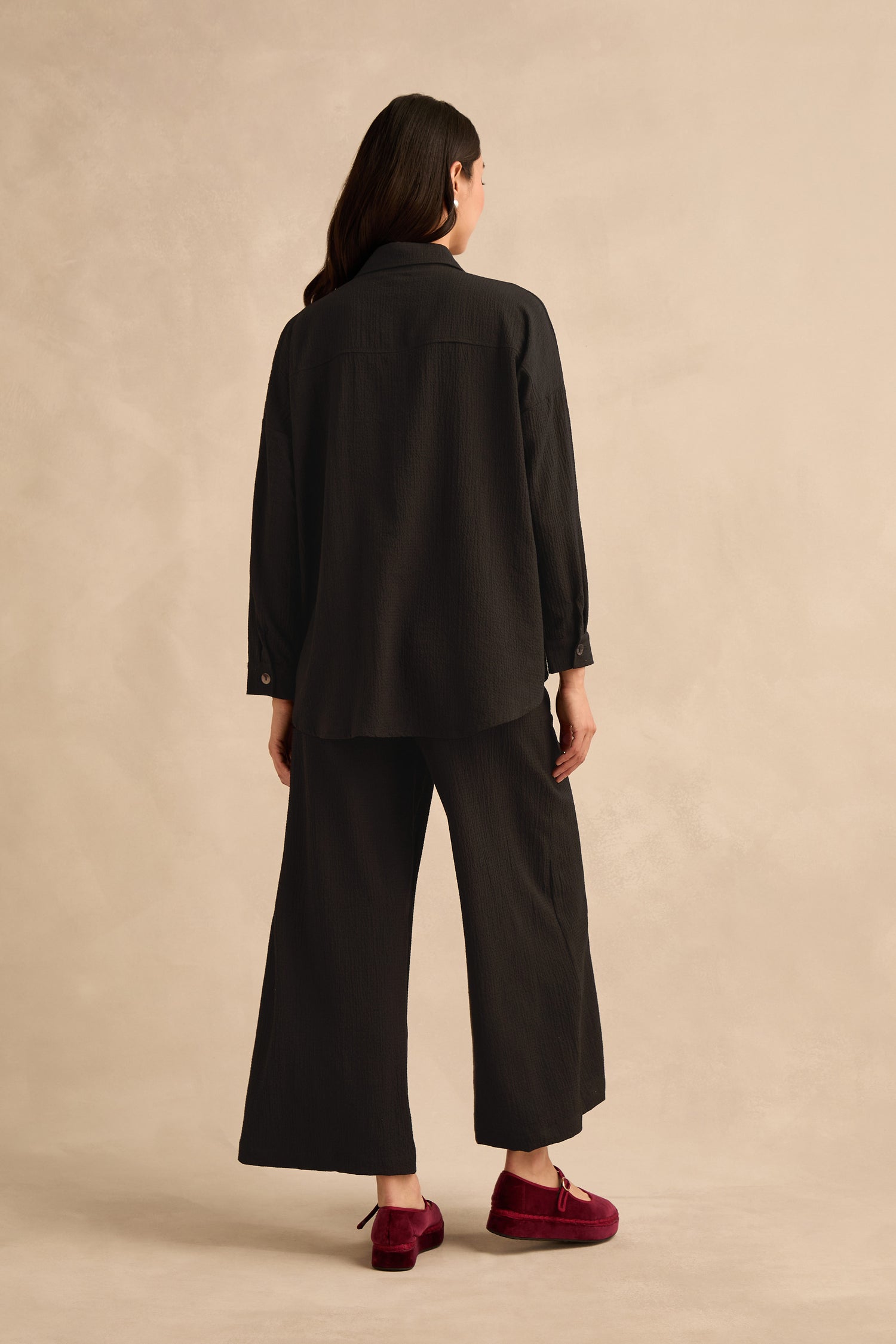 Wearing the Mini Crinkle Viscose Boxy Shirt, a person faces away from the camera, styled in wide-leg pants and burgundy shoes against a beige background.