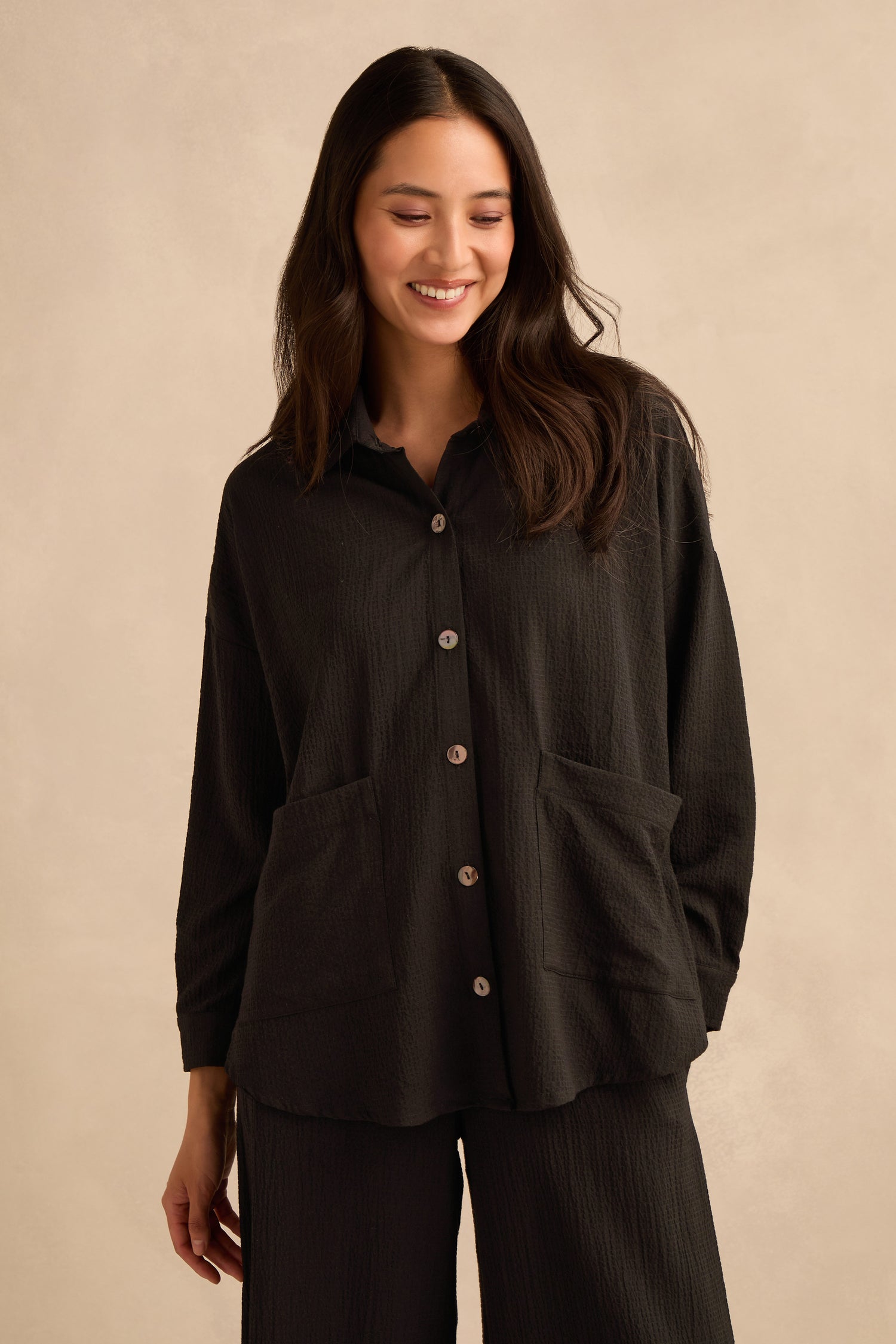 A woman wears the Mini Crinkle Viscose Boxy Shirt—an oversized, textured black button-up with large front pockets—paired with matching black pants, standing against a plain beige background.