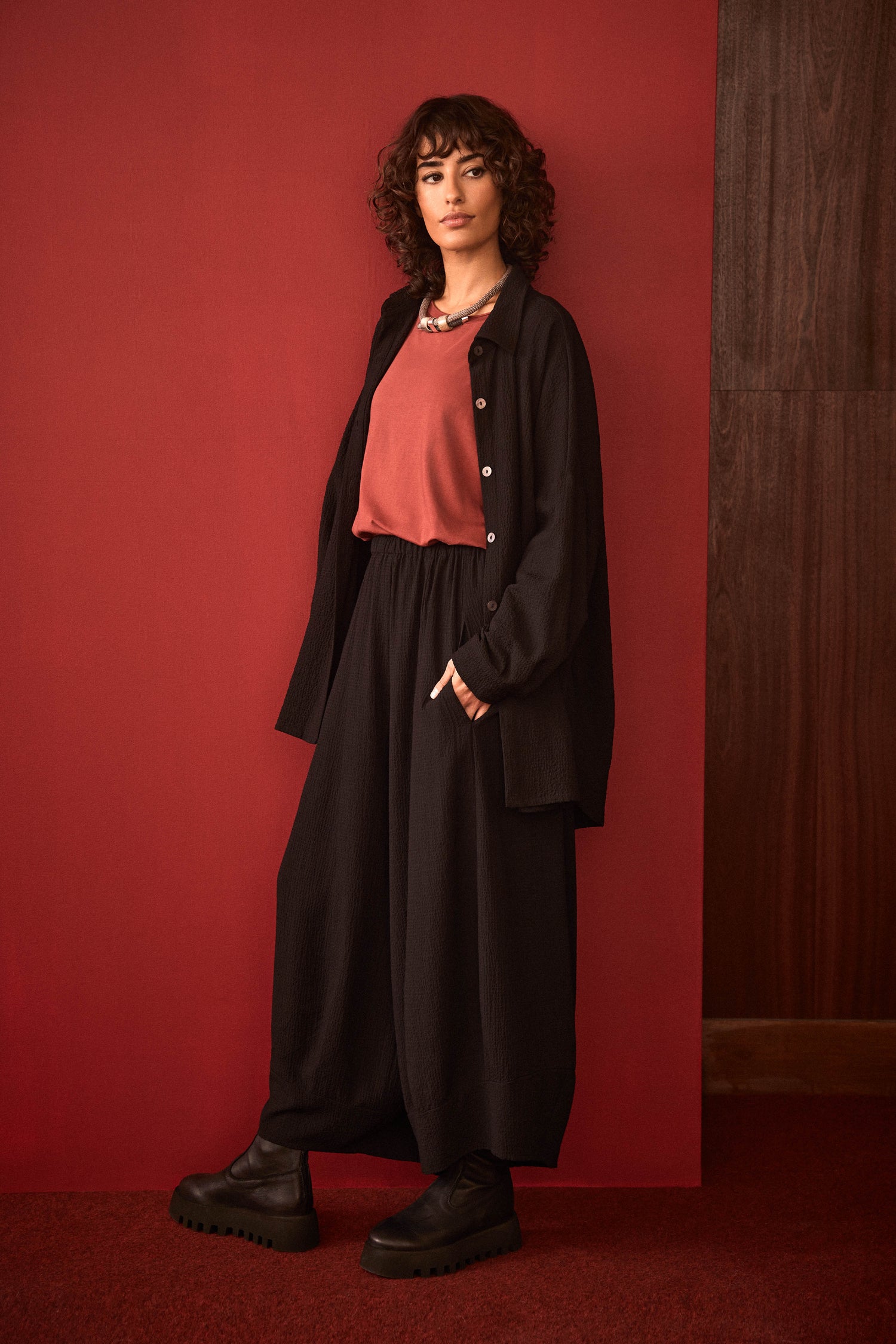 A person with curly hair stands against a red background, wearing the Fine Crinkle Viscose Boxy Shirt in black, a coral top, chunky black shoes, and a necklace.