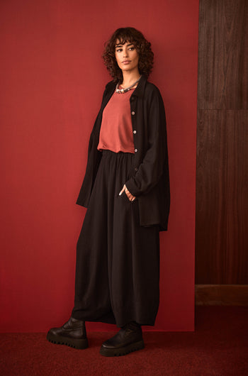 A person with curly hair stands against a red background, wearing the Fine Crinkle Viscose Boxy Shirt in black, a coral top, chunky black shoes, and a necklace.