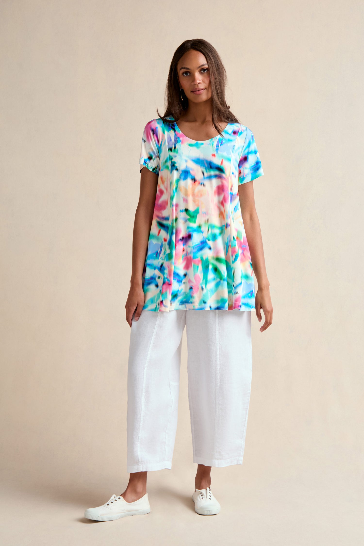 A woman stands against a neutral background wearing the Petal Haze Jersey Tunic—a relaxed-fit, colorful jersey tunic with a subtle floral print—paired with white wide-leg pants and white sneakers.