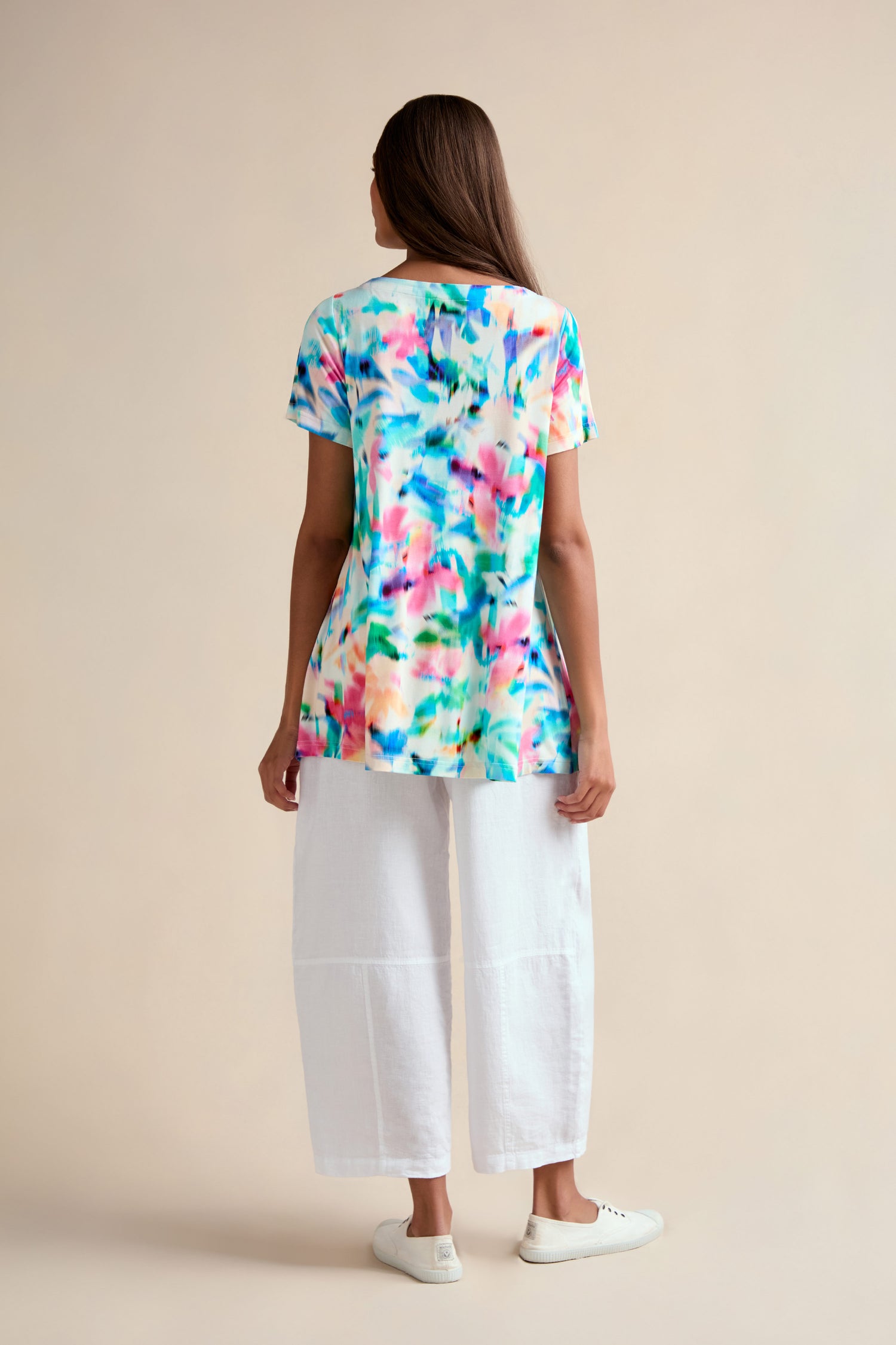 A woman faces away wearing the Petal Haze Jersey Tunic—a relaxed fit, colorful short-sleeve top with a watercolor pattern—paired with white wide-leg pants and white shoes against a plain background.