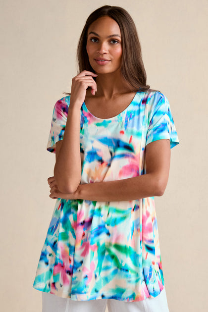 A woman wears the Petal Haze Jersey Tunic, featuring a short-sleeved, relaxed fit and a vibrant abstract print, standing against a plain beige background.