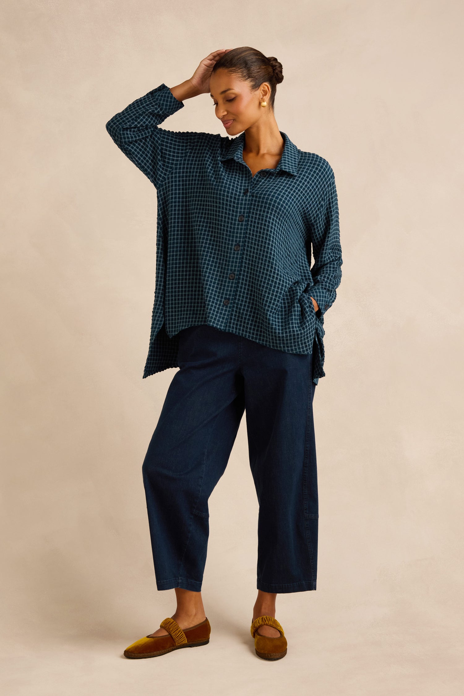 A person stands against a plain beige background, wearing the Seersucker Squares Relaxed Shirt with a boxy silhouette, paired with wide-leg dark blue pants and brown shoes—one hand touching their head and the other in their pocket.